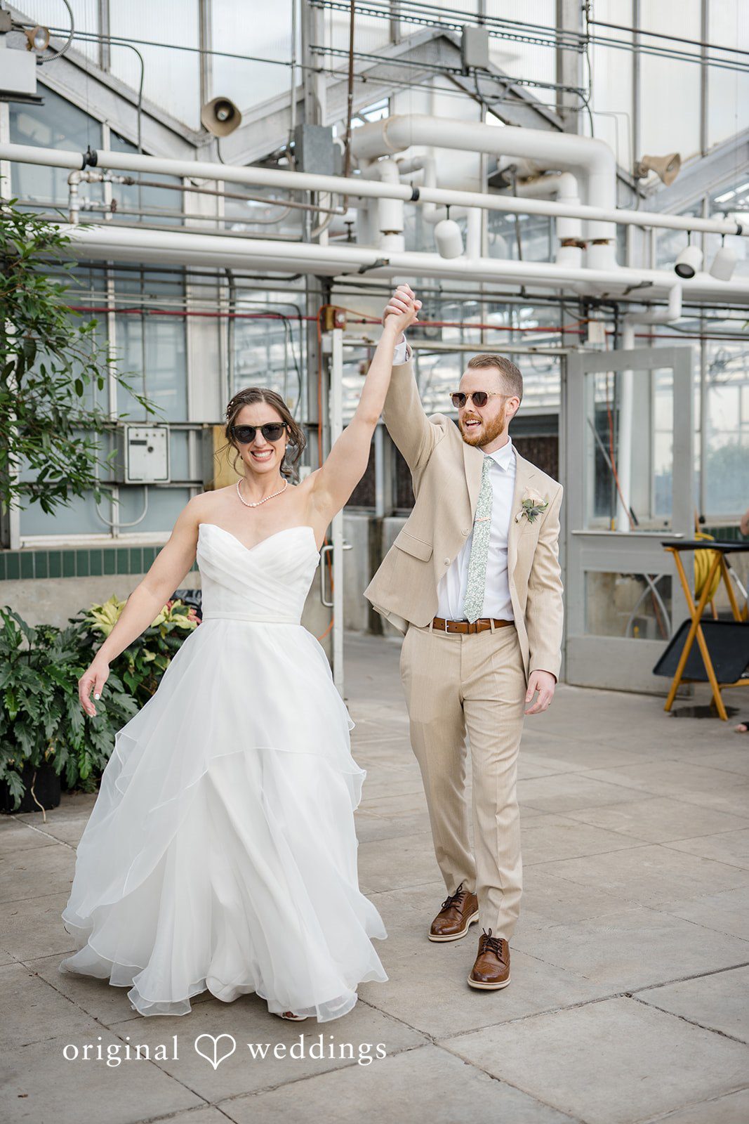 Emily + Maxwell A bride and groom celebrating with raised arms inside a spacious glass greenhouse structure.