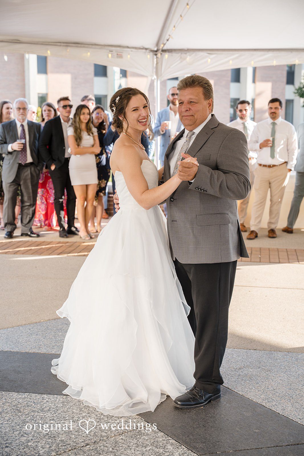 Emily + Maxwell A bride in a white gown dancing with a man in a grey suit jacket as wedding guests watch.