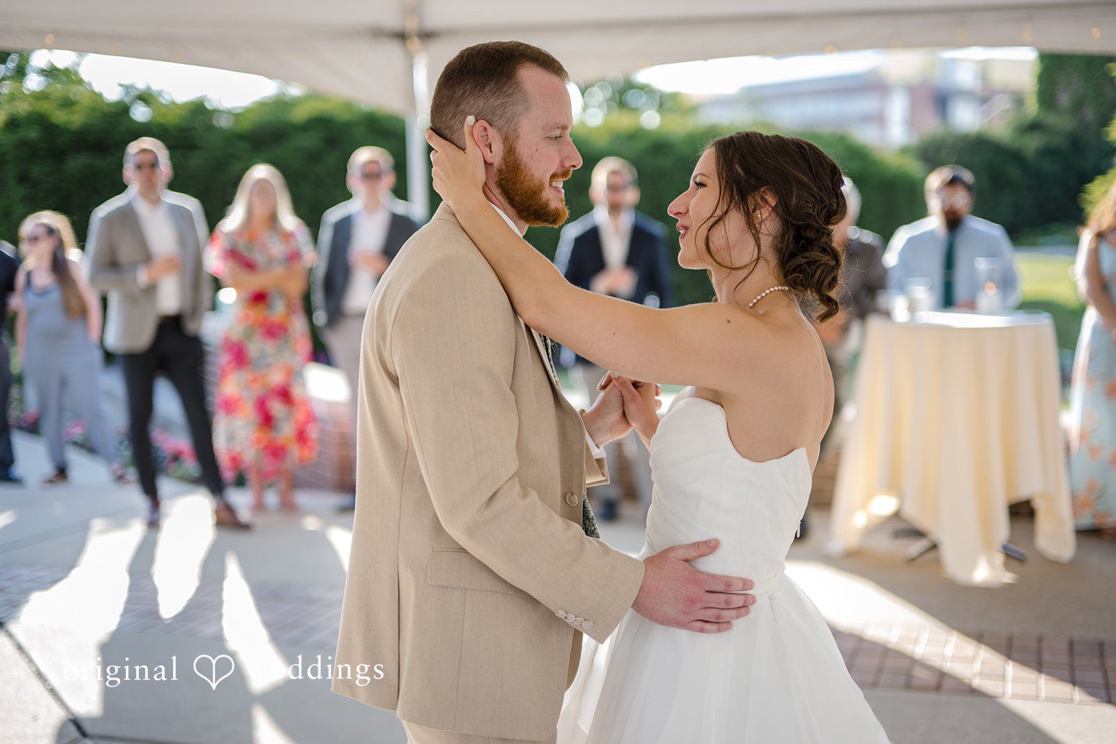 Emily + Maxwell A bride and groom embracing and looking at each other during their first dance while guests watch in the background.