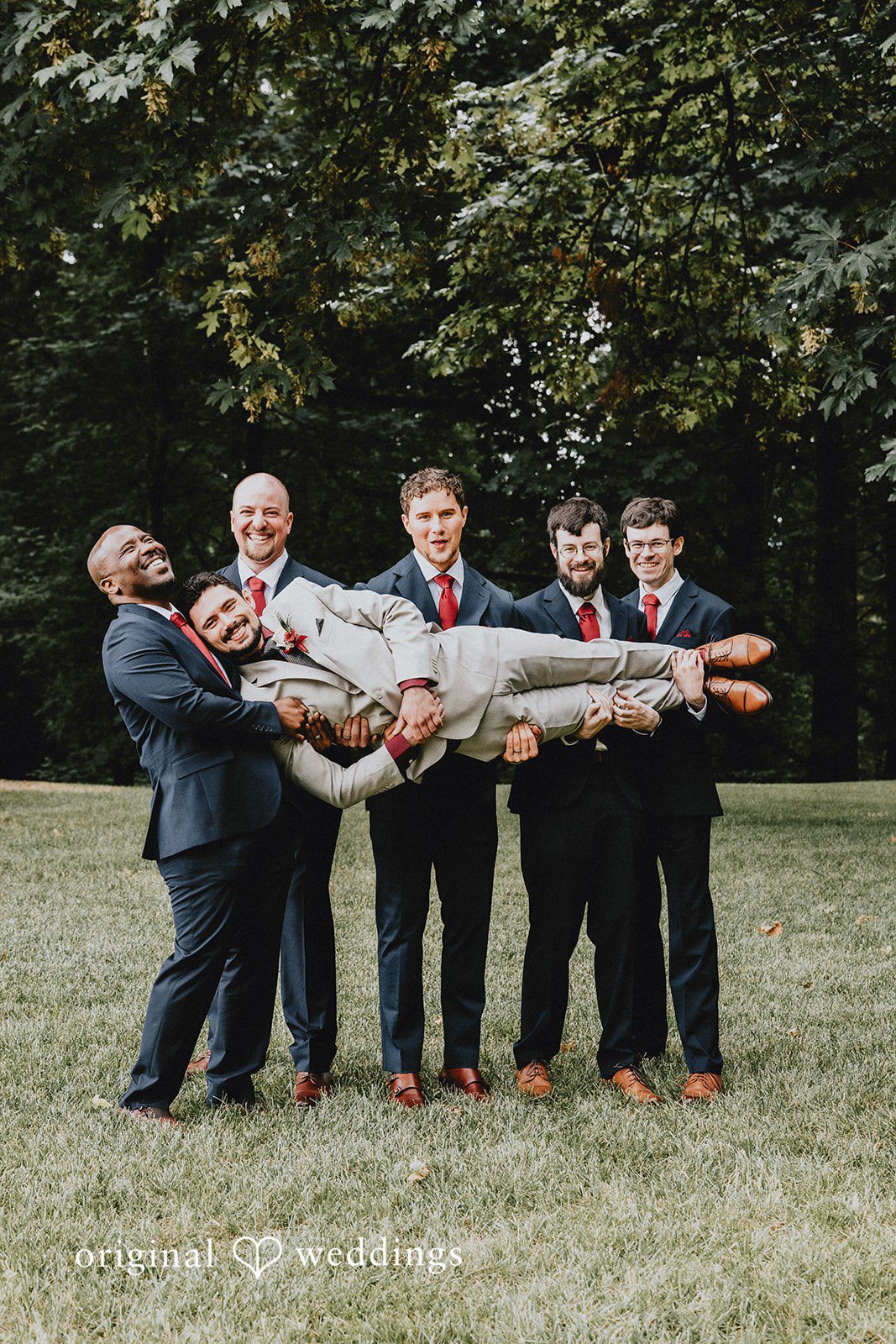 Catherine + Gabriel Groomsmen playfully lifting the groom horizontally in the air on a lawn.