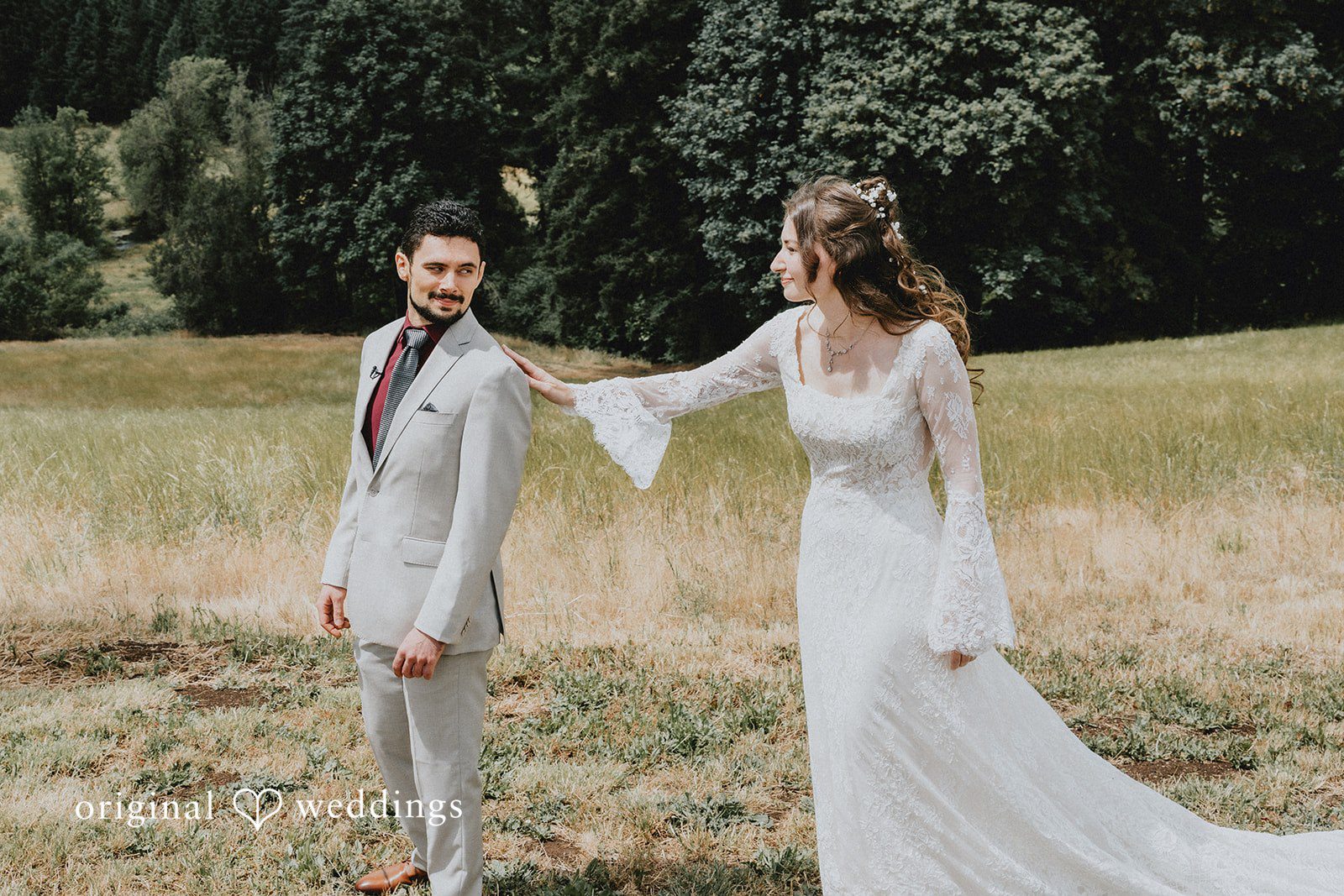 Catherine + Gabriel Bride in a long-sleeved lace gown tapping a groom in a grey suit on the shoulder during a first look in a grassy field.