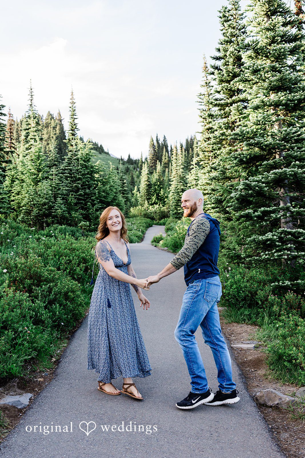 Megan + Kevin A man and a woman hold hands and face each other on an asphalt path cutting through a pine forest.