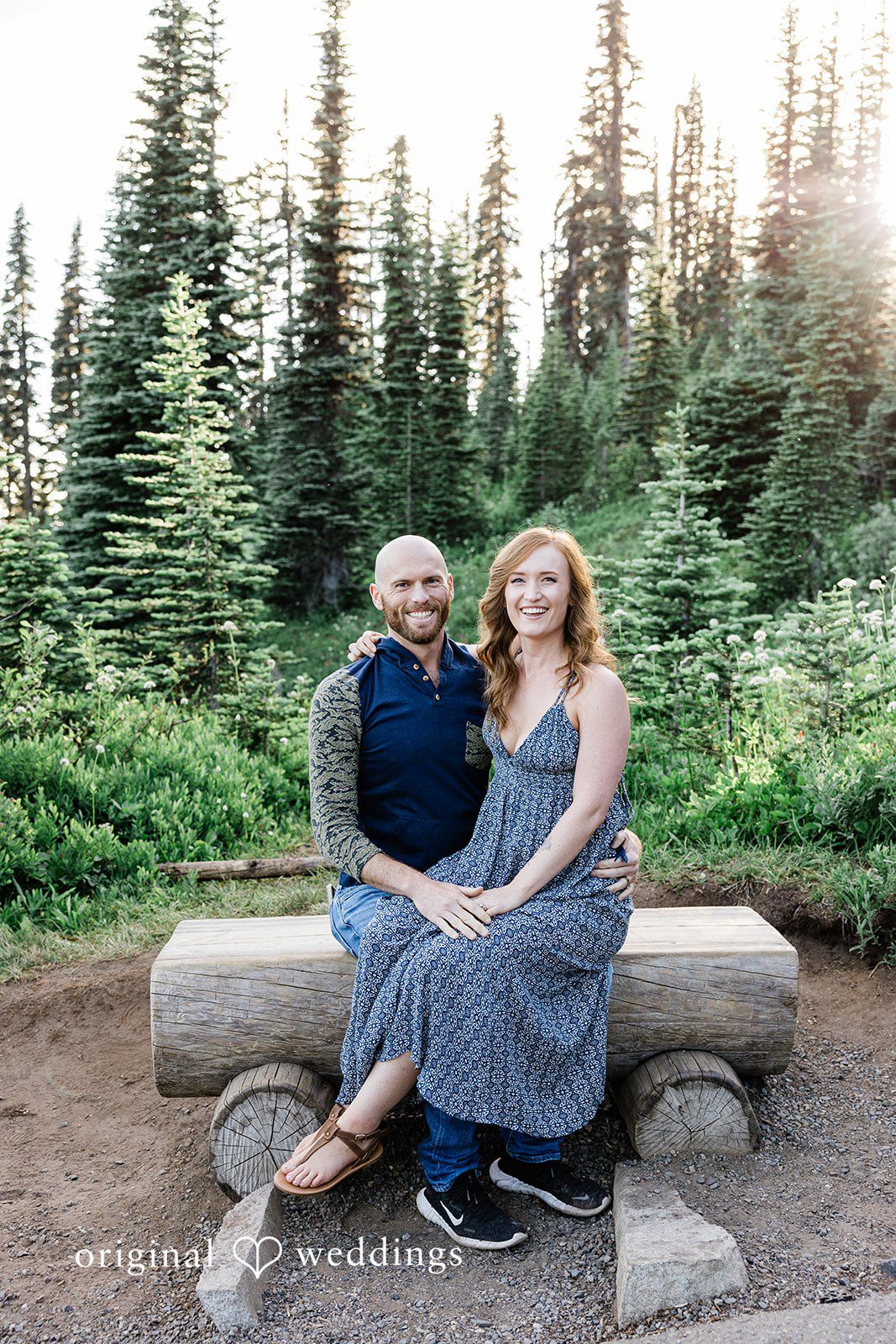 Megan + Kevin A man and a woman sit together on a rustic wooden bench surrounded by evergreen trees.