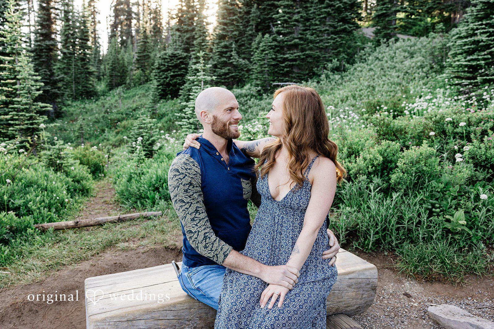 Megan + Kevin A man and a woman look at each other while sit on a rustic wooden bench with tall trees.