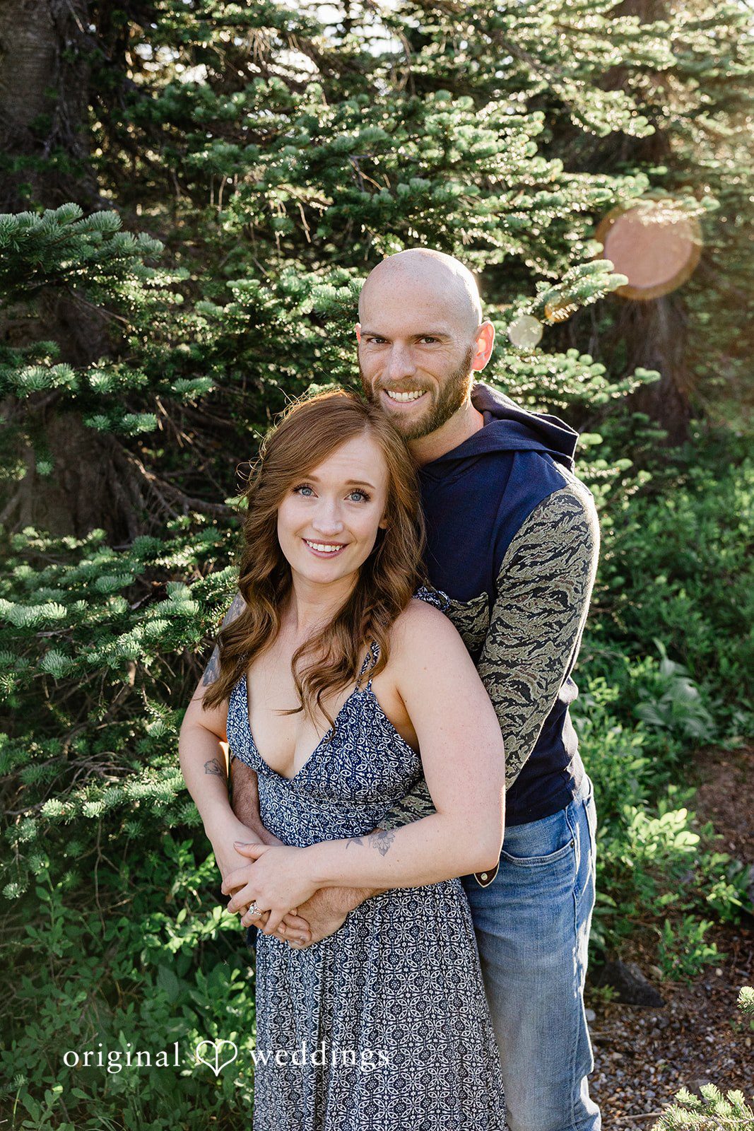Megan + Kevin A man stands behind a smiling woman in a blue dress with a pine tree background.