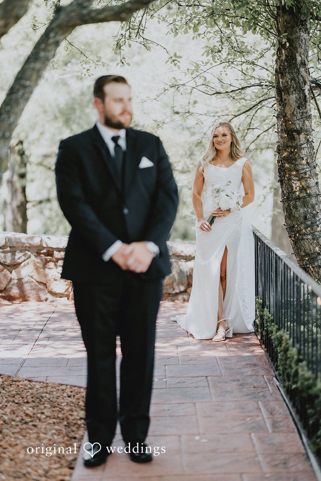 Groom standing in foreground while bride walks down path.