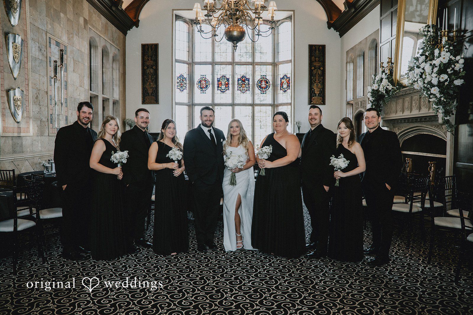 Wedding party in black attire standing inside a grand hall.