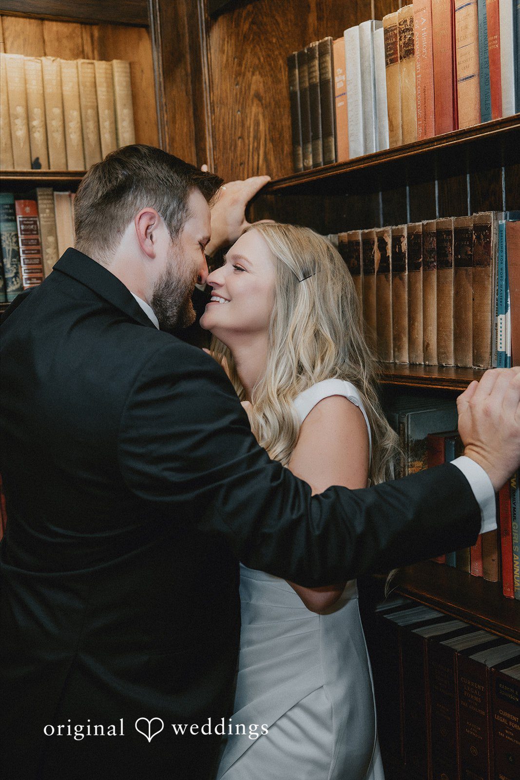 Groom leaning in to embrace the bride in front of bookshelves.