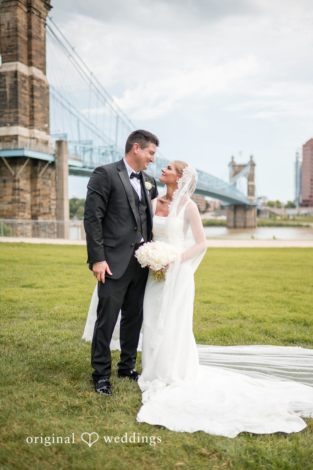 Bride and groom smiling together on a grassy hill with a bridge background.