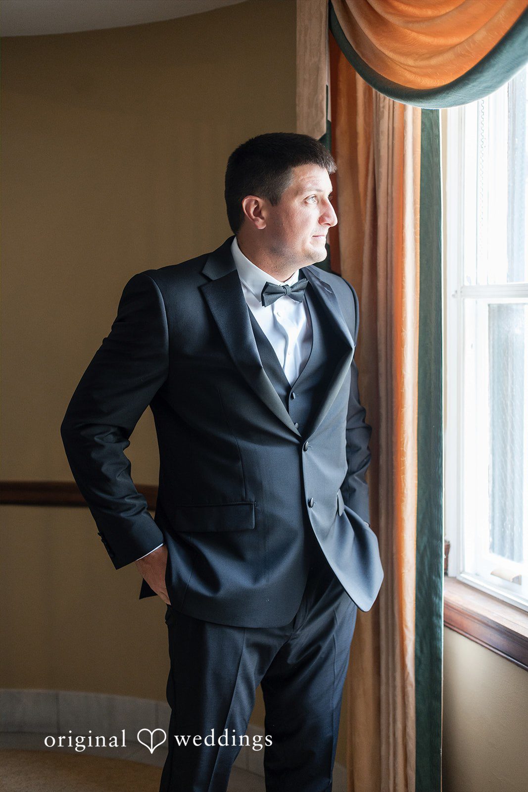 Groom standing alone by a window looking off to the side.