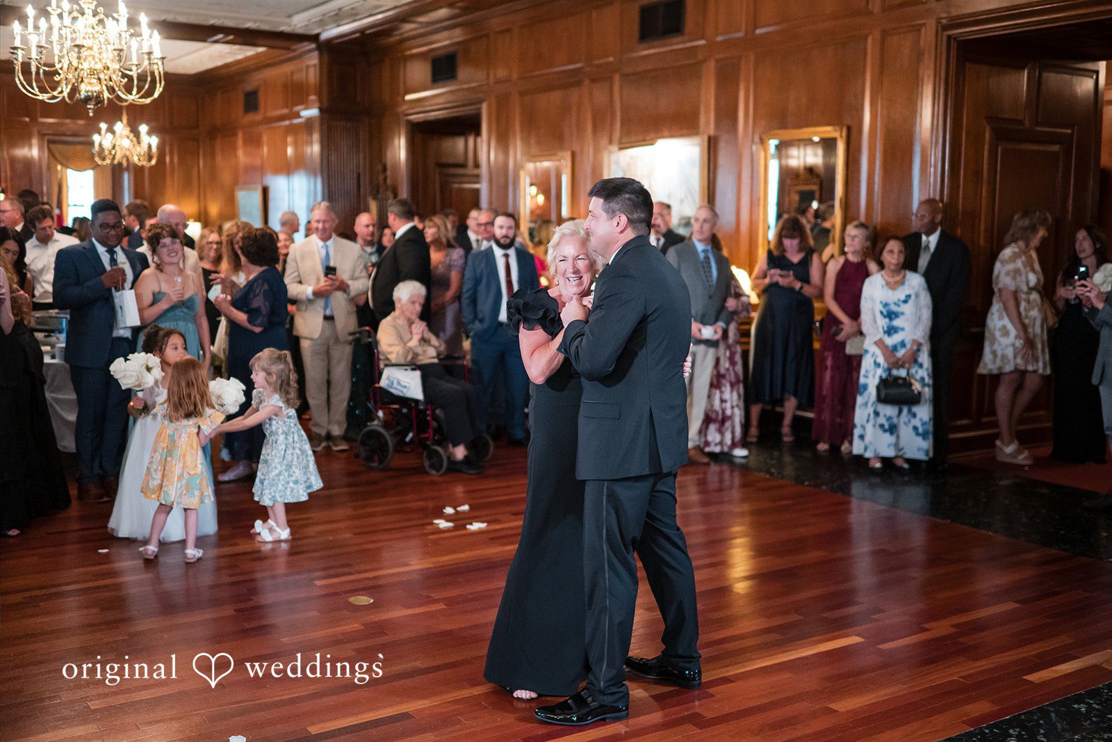 A smiling woman in a black gown and a man in a black tuxedo dance together on a wooden floor.