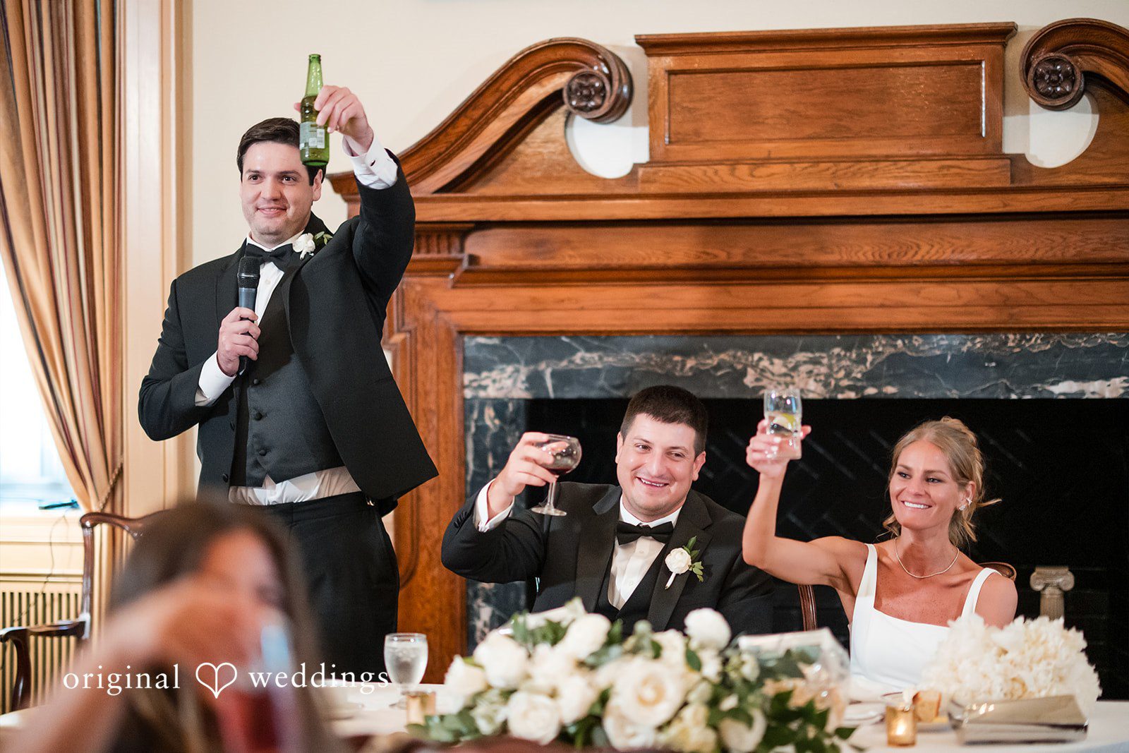 A man in a tuxedo raises a beer bottle and speaks into a microphone while a seated bride and groom raise their glasses in a toast.