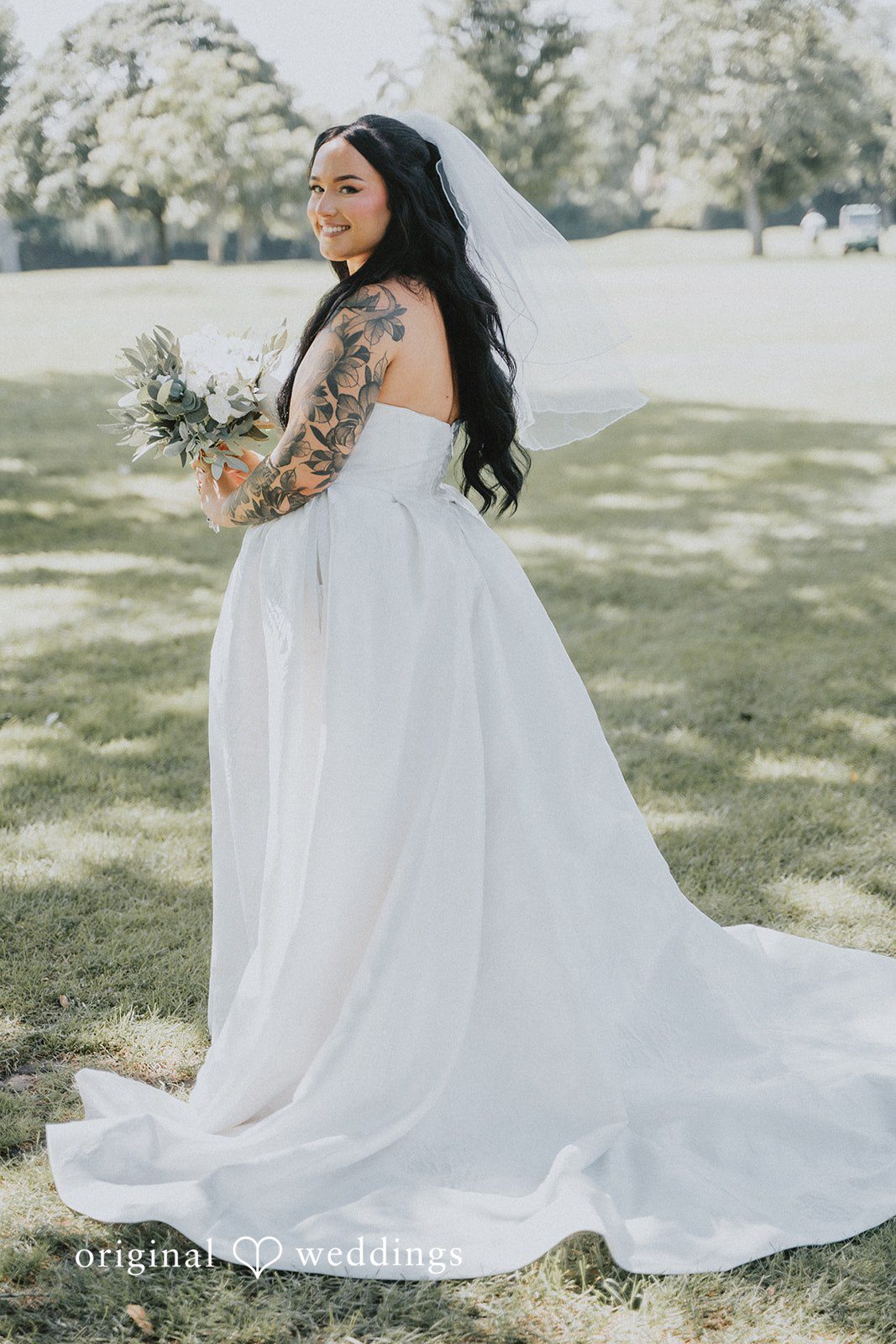 Carly + Dillon A bride in a white gown posing from behind while holding a bouquet on a grassy lawn.