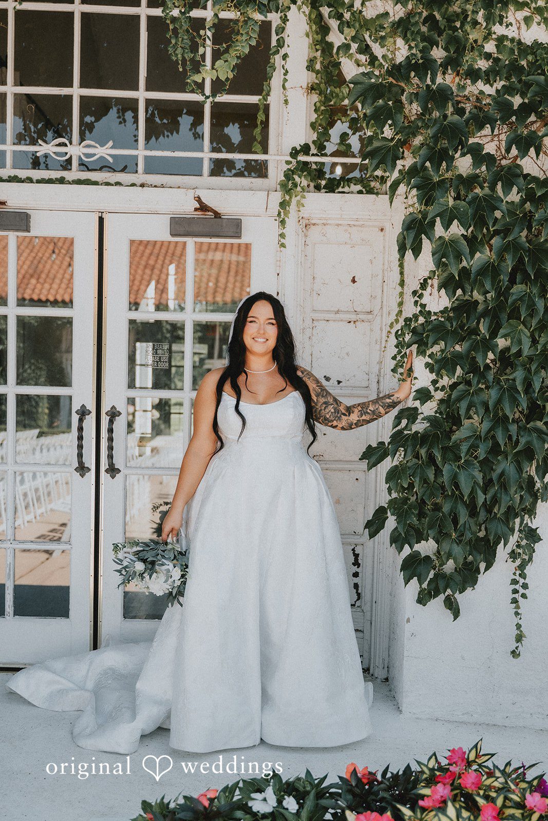 Carly + Dillon A bride in a white wedding dress standing near an arched doorway with greenery.