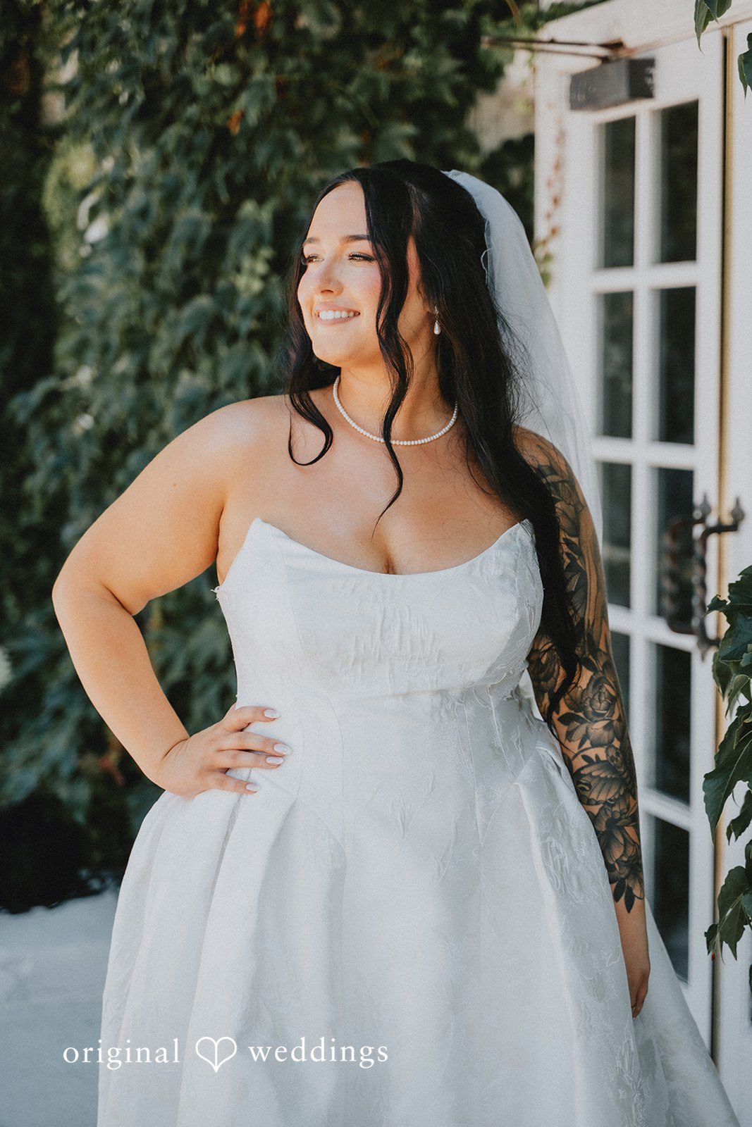 Carly + Dillon A bride in a white wedding gown and veil smiling while posing next to a building.