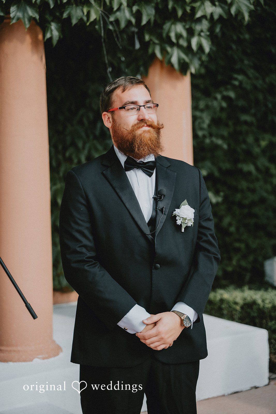 Carly + Dillon A groom in a black tuxedo and glasses posing for a portrait against a brick wall.