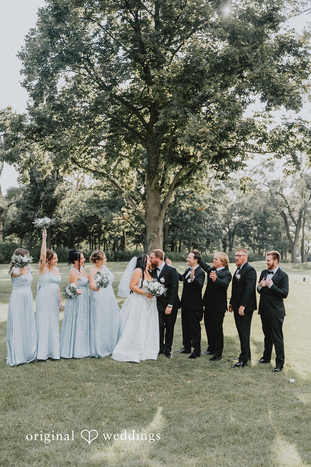 Carly + Dillon A wedding party with bridesmaids and groomsmen posing together under a large tree.