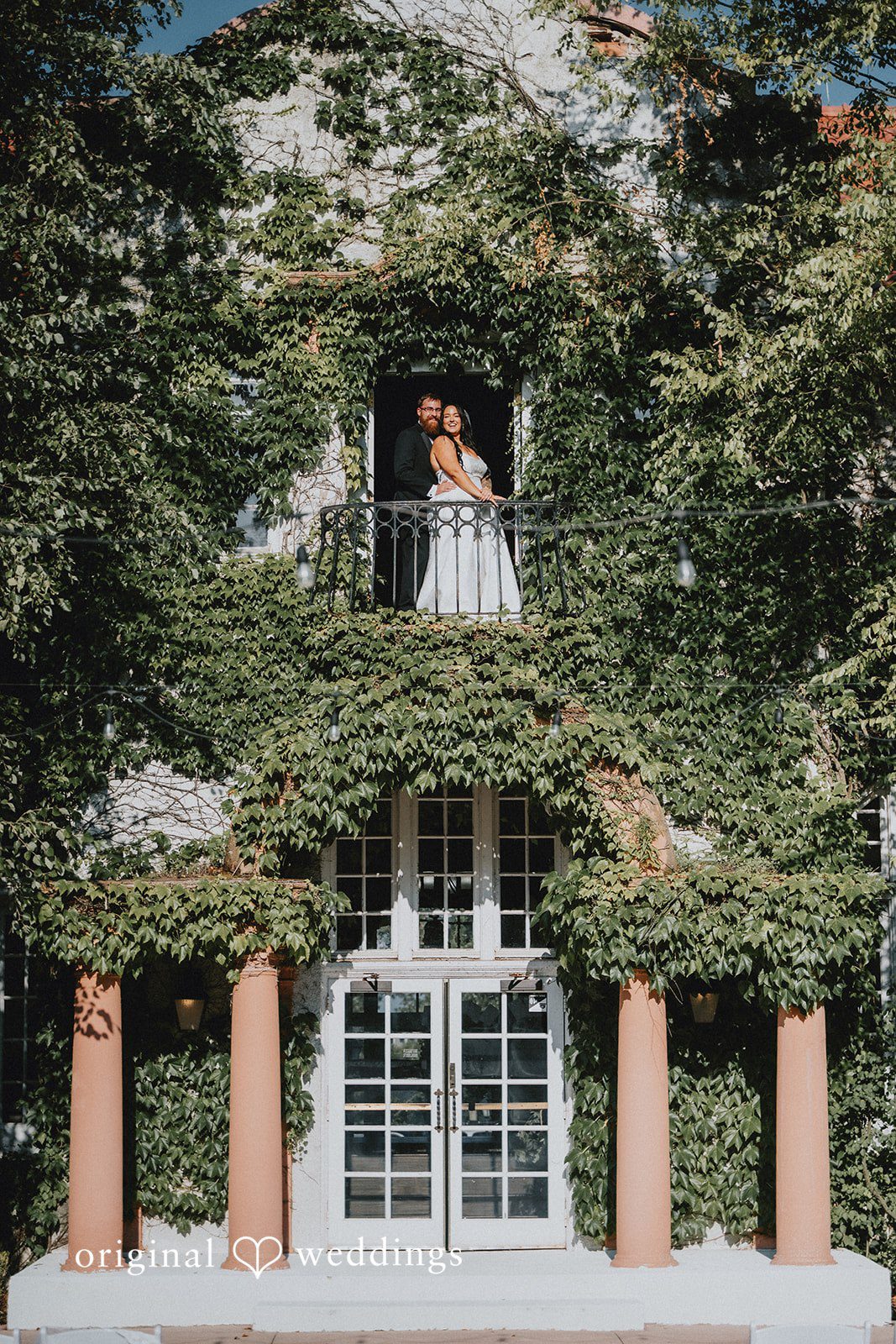 Carly + Dillon A bride looking down from a high balcony covered in lush green ivy.