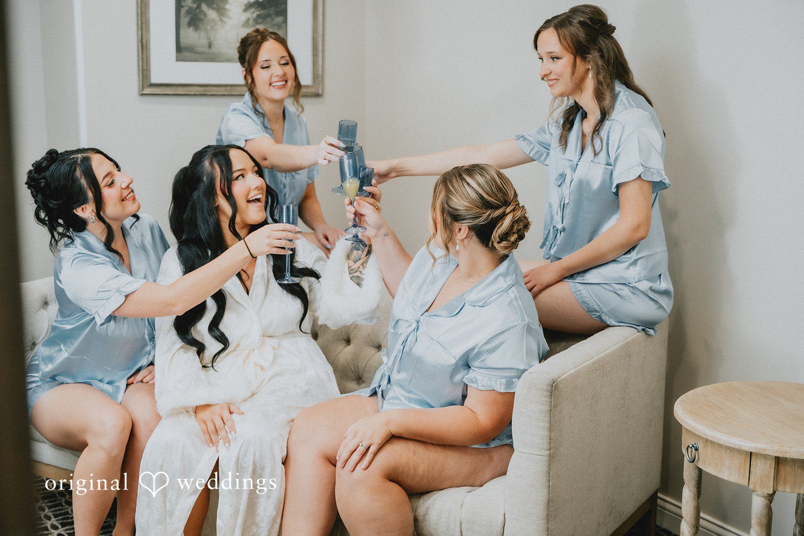 Carly + Dillon A bride and her bridesmaids in matching blue silk pajamas sitting together on a couch.