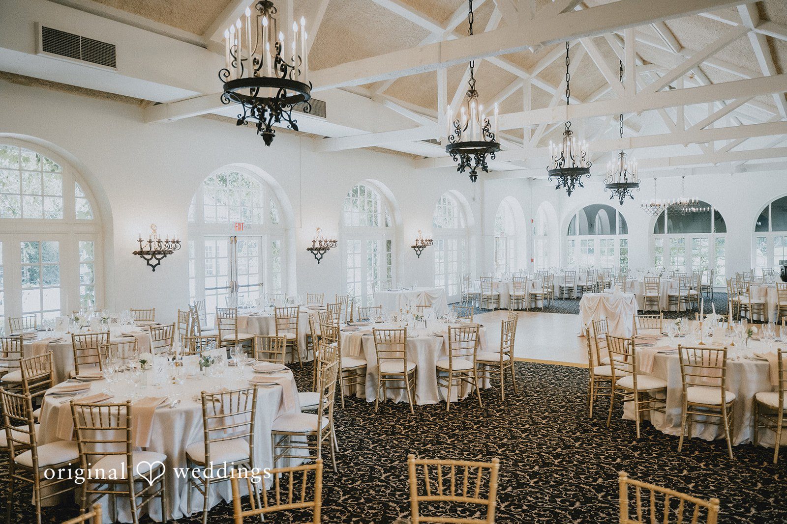 Carly + Dillon A large, brightly lit reception hall filled with round dining tables and chairs.