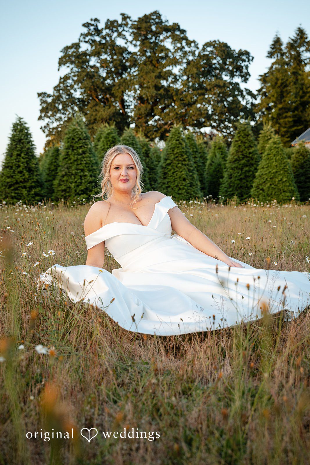 Victoria + Skyler Bride in a white wedding gown sitting on a grassy field.