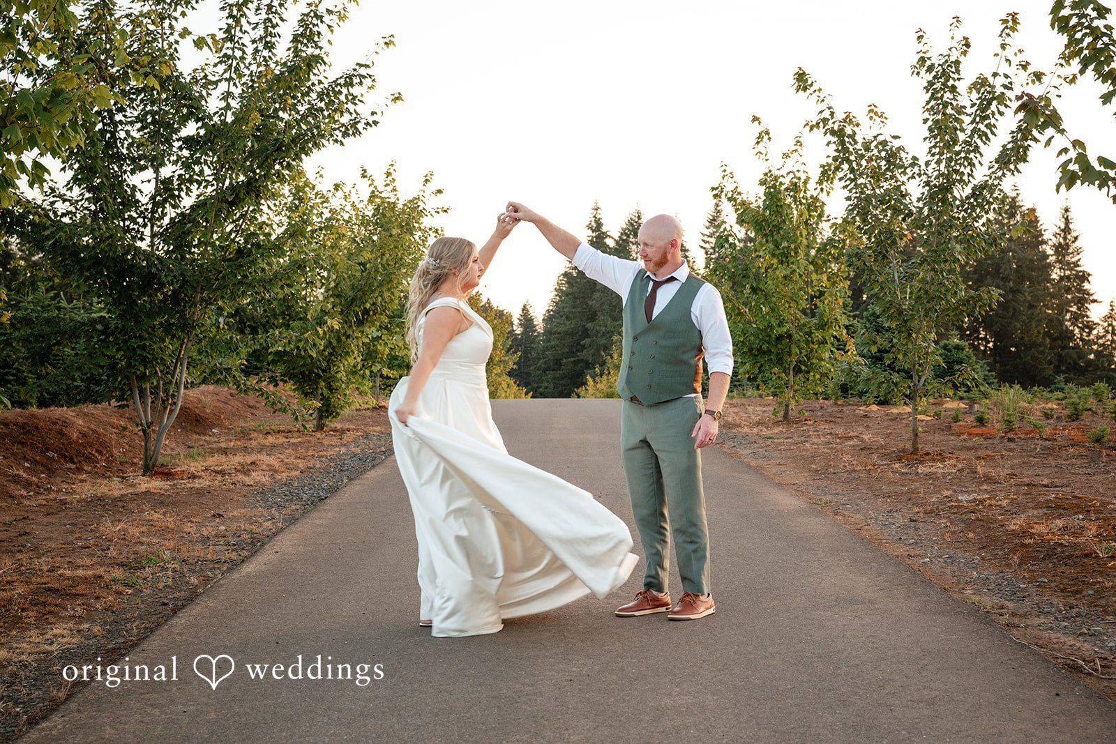 Victoria + Skyler Bride and groom dancing together on a dirt path lined with trees.