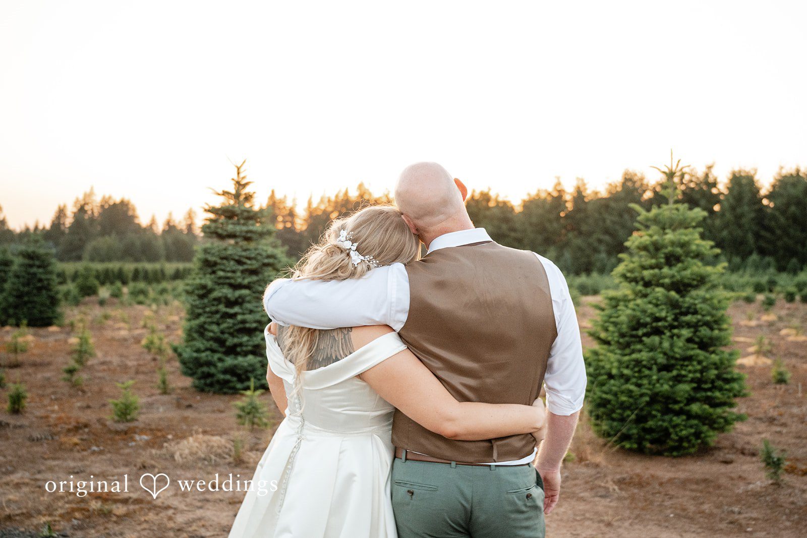 Victoria + Skyler Bride and groom standing with their arms around each other in a field of trees.