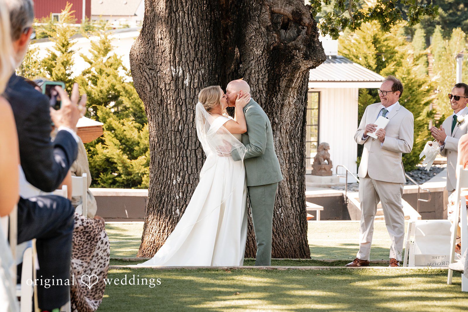 Victoria + Skyler Bride and groom kissing under a large tree during their ceremony.