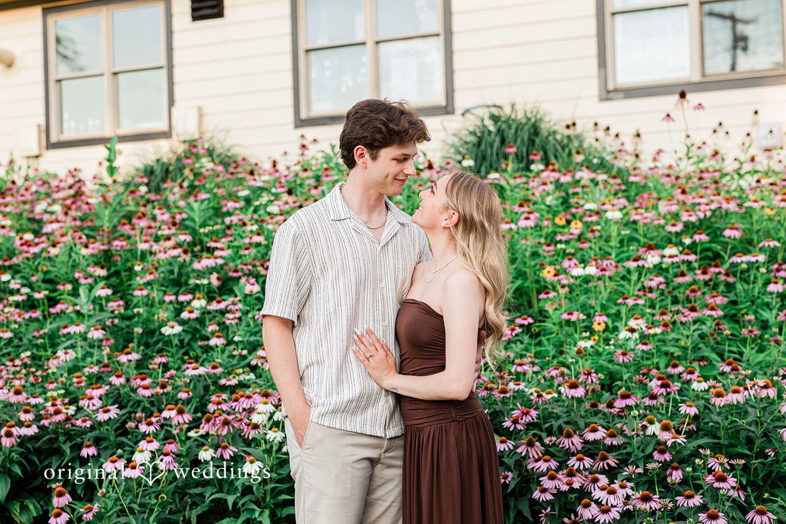 Sara + Jack A couple embracing closely in front of a building with a flower-filled garden.