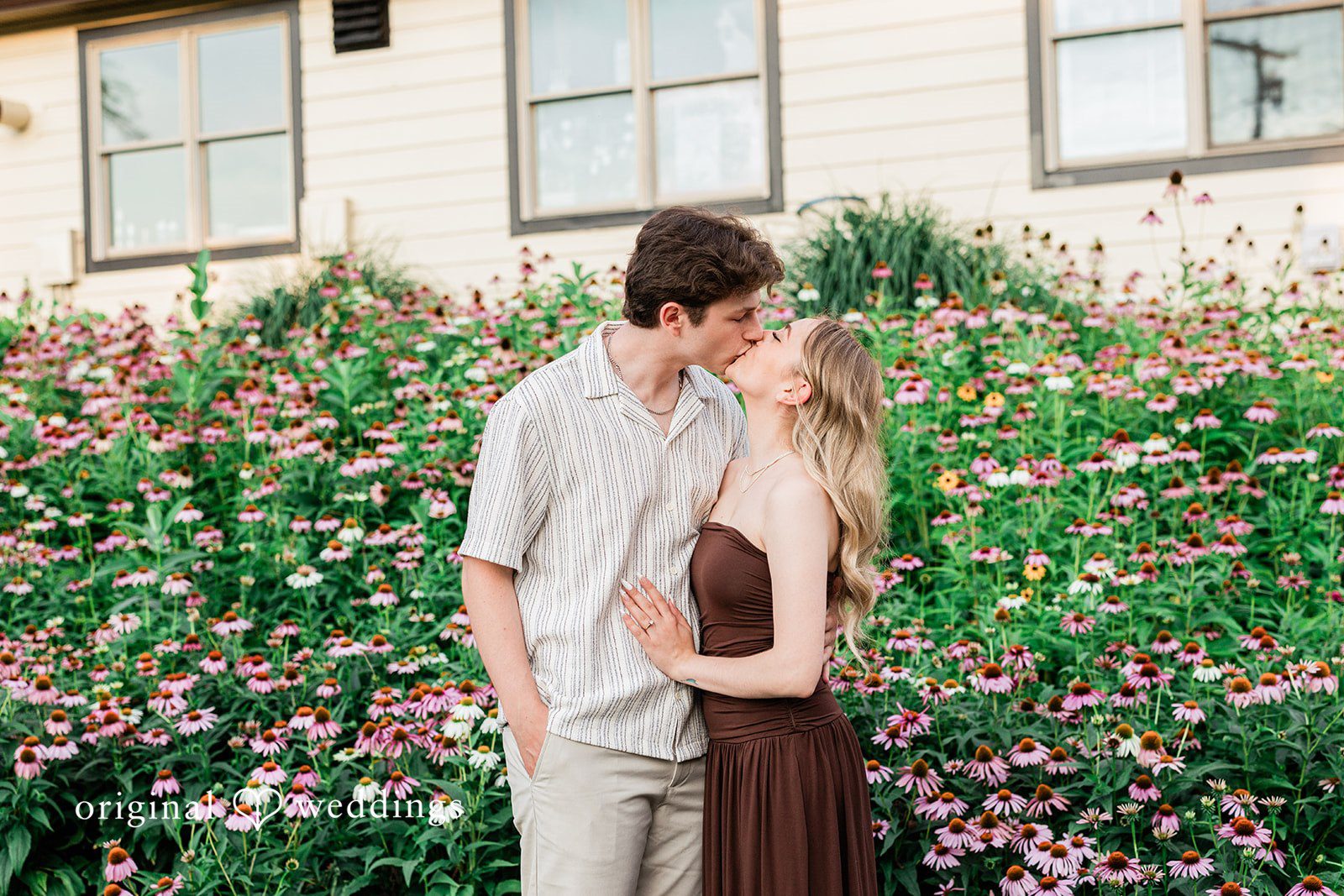 Sara + Jack A couple embracing closely in front of a building with a flower-filled garden.