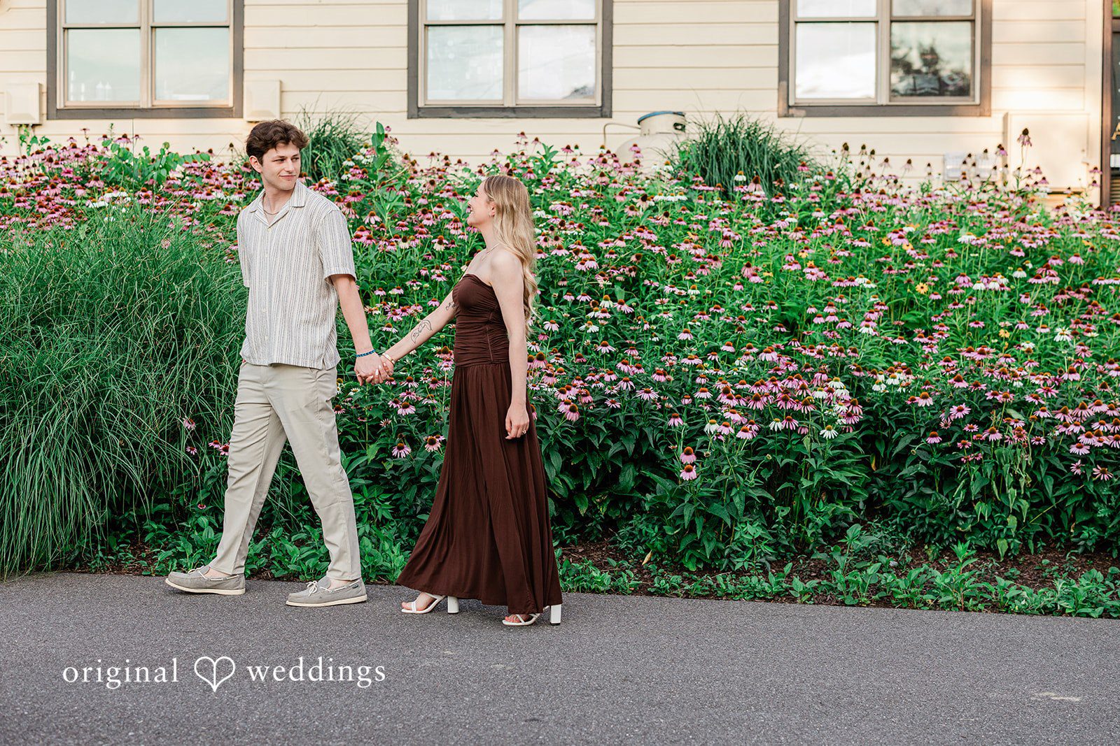 Sara + Jack A couple holding hands while walking on a paved path next to a flower garden.