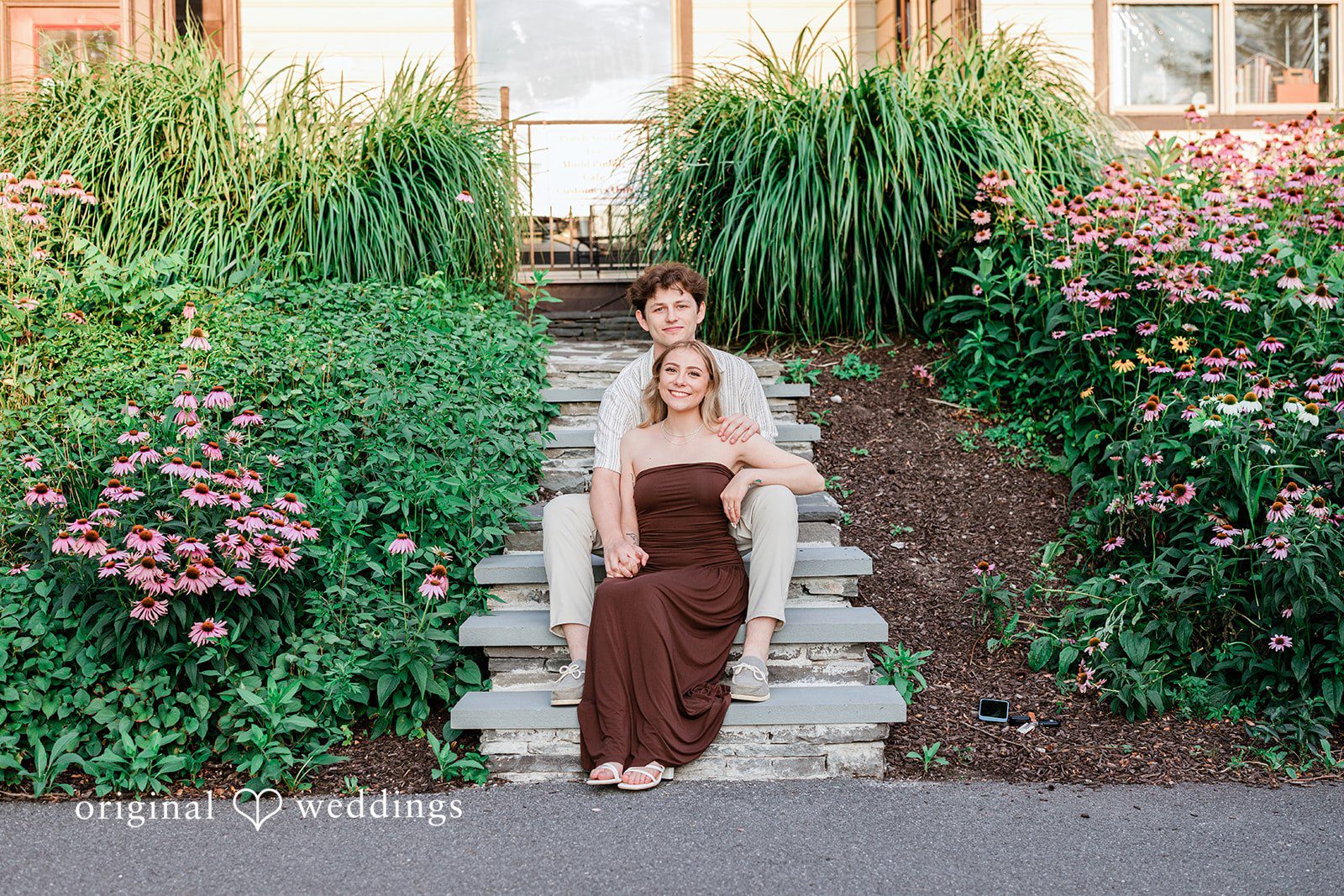 Sara + Jack A couple posing together on stone steps surrounded by lush green bushes.