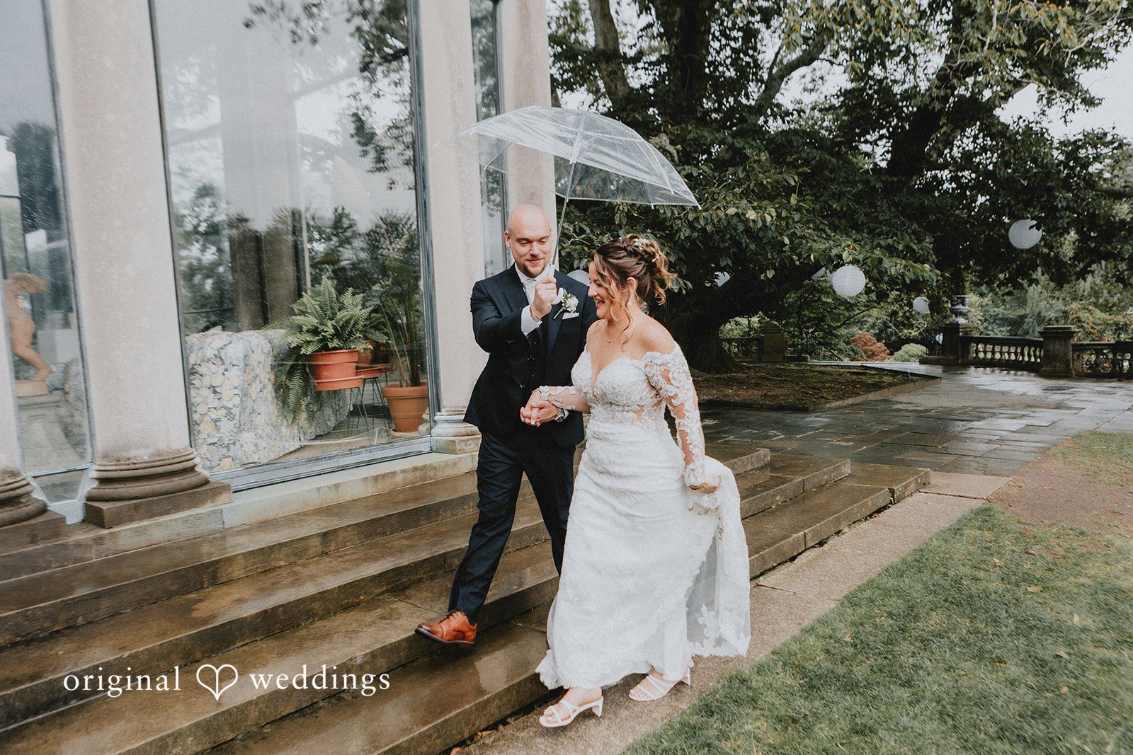 Samantha + Michael Bride and groom standing under an umbrella on a rainy path.