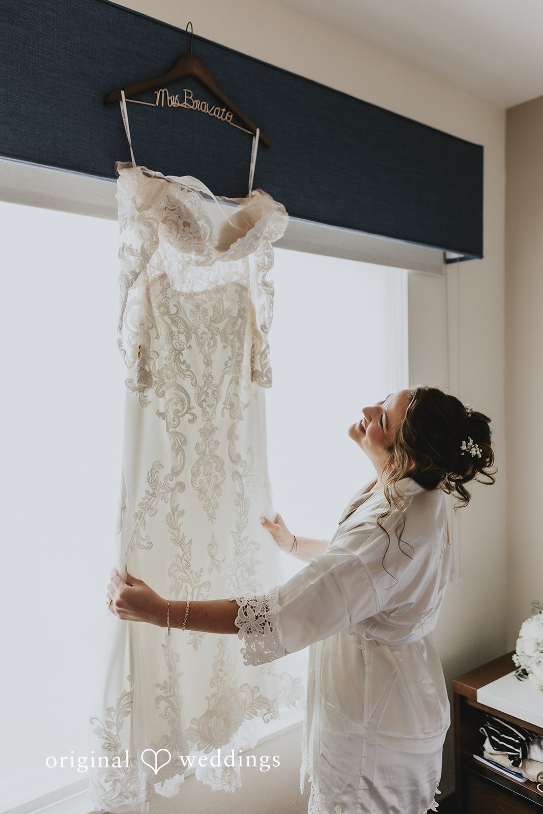 Samantha + Michael Bride looking up at her wedding dress hanging near a window.
