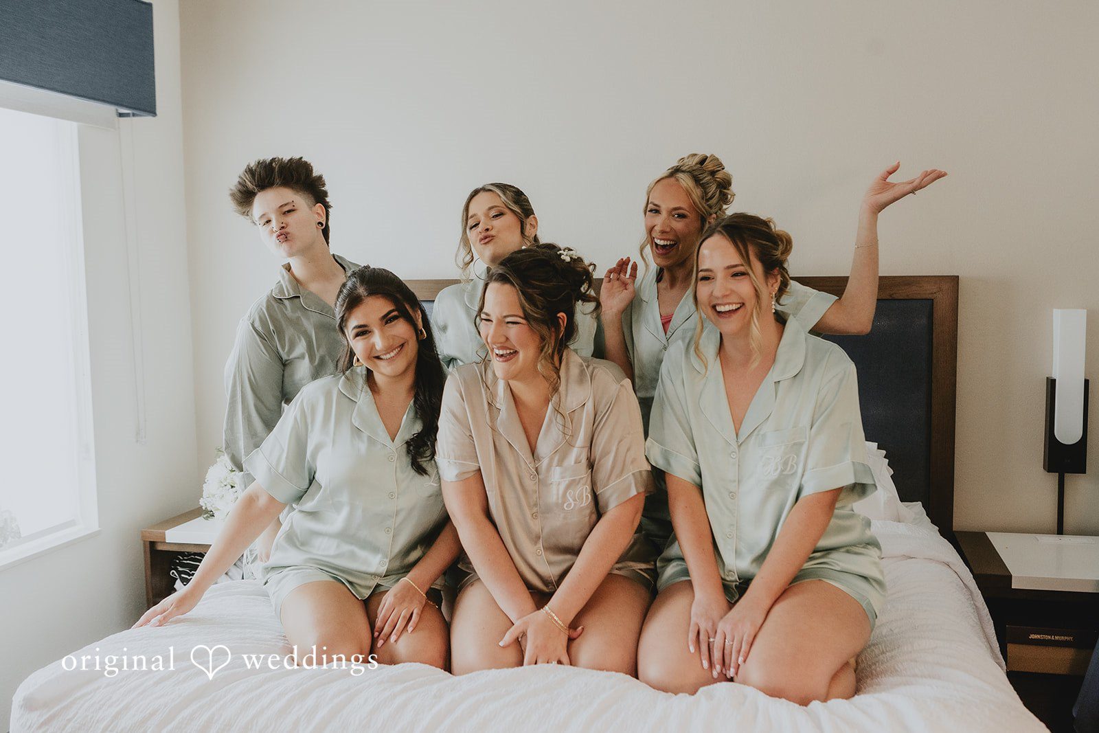 Samantha + Michael Group of bridesmaids in silk robes laughing together on a bed.