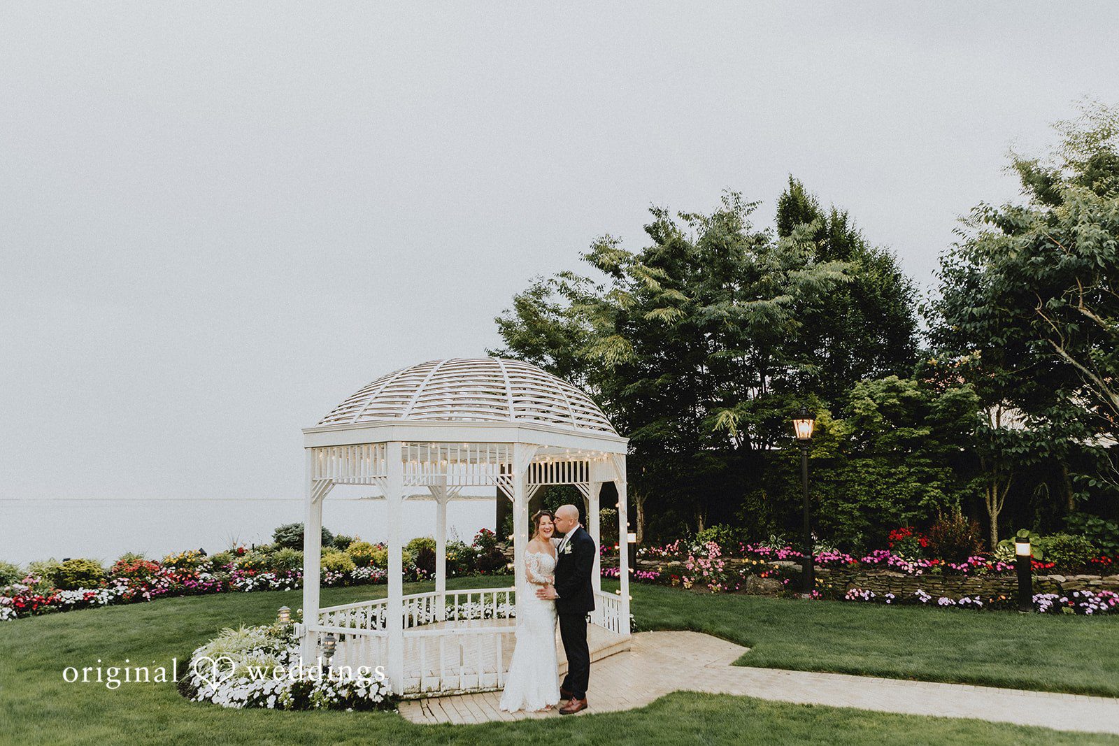 Samantha + Michael Distant view of a bride and groom standing by a white garden gazebo.
