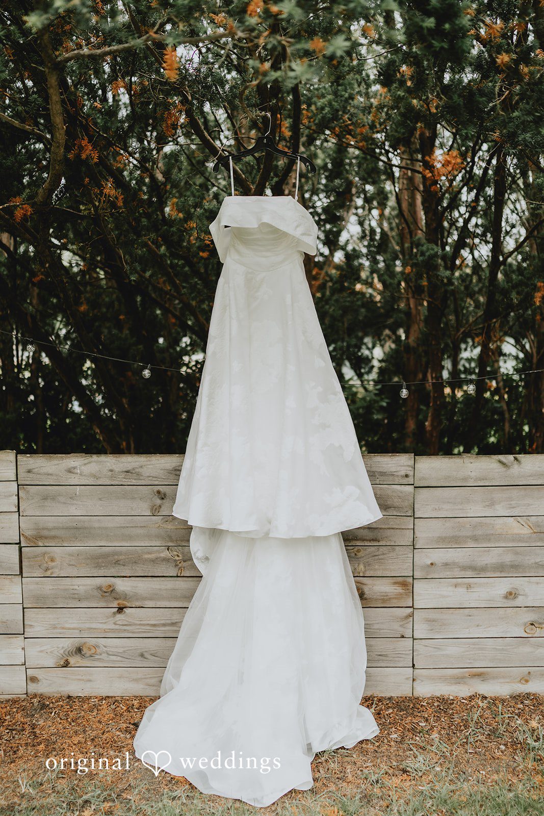 Brandon + Natalie A white tiered wedding dress hanging against a rustic wooden fence.
