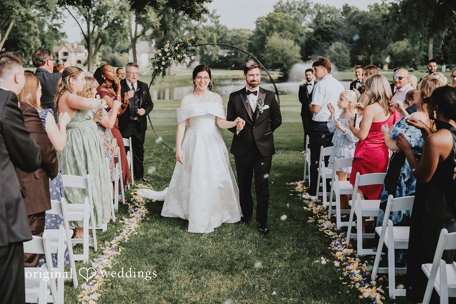 Brandon + Natalie A bride and groom walking down a grassy aisle lined with seated wedding guests.
