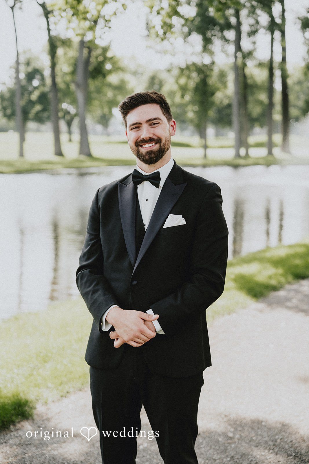 Brandon + Natalie A groom in a black tuxedo smiling while posing by a lake.