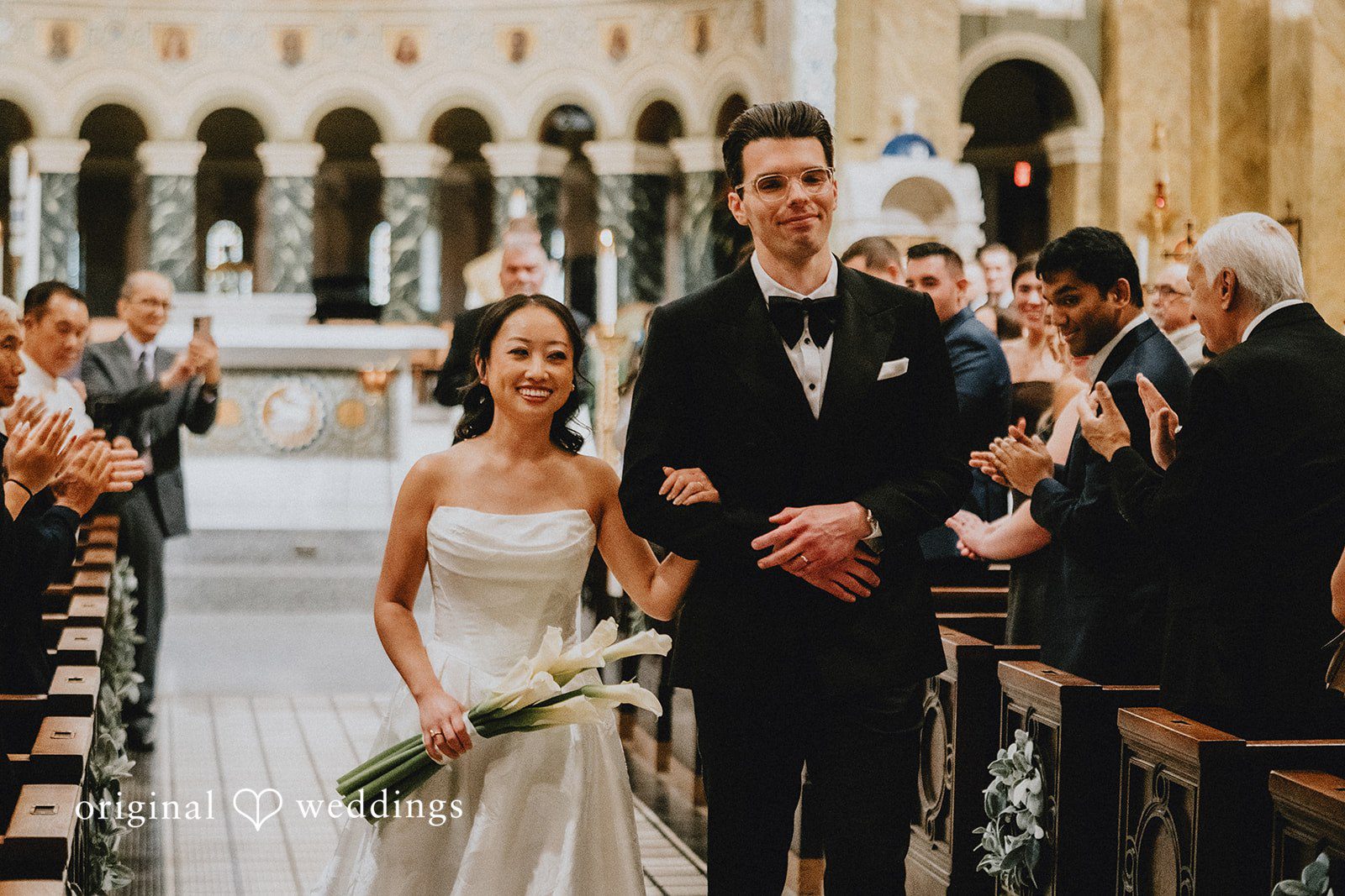 Aleksandar + Anna A newly married bride and groom walking down the aisle together smiling.