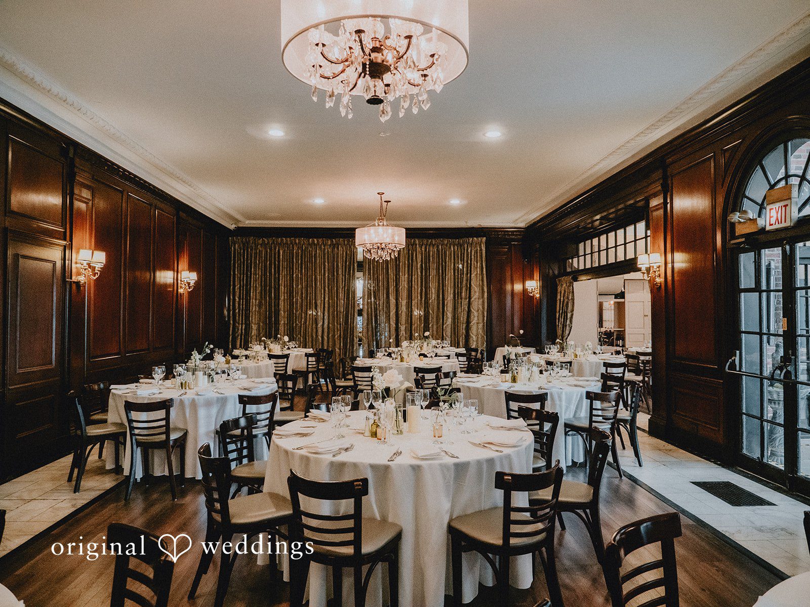 Aleksandar + Anna An elegant reception hall set up with round tables, white tablecloths, and lit candles.