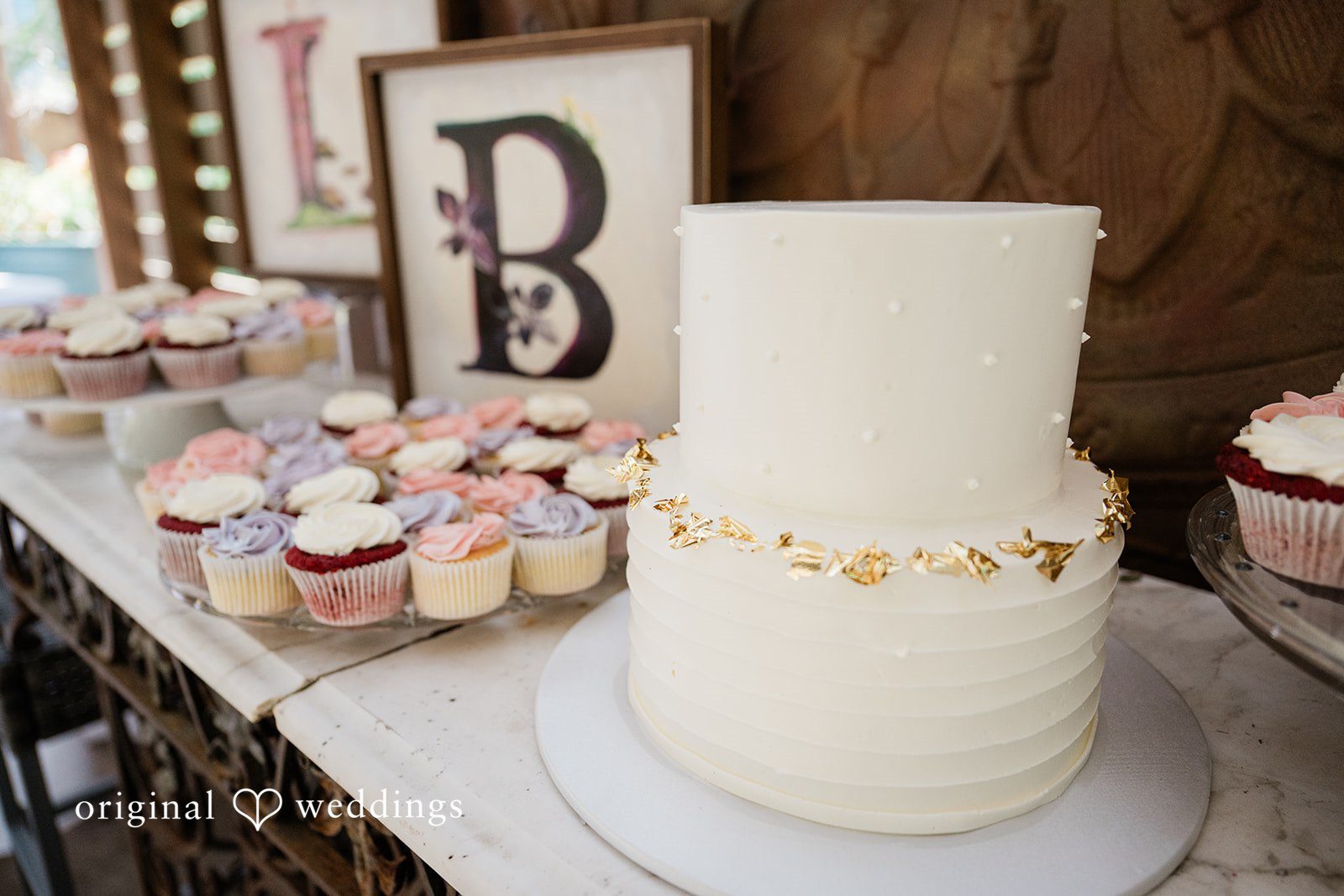 Lauren + Brinton A multi-tier white wedding cake decorated with gold leaf accents on a dessert table.