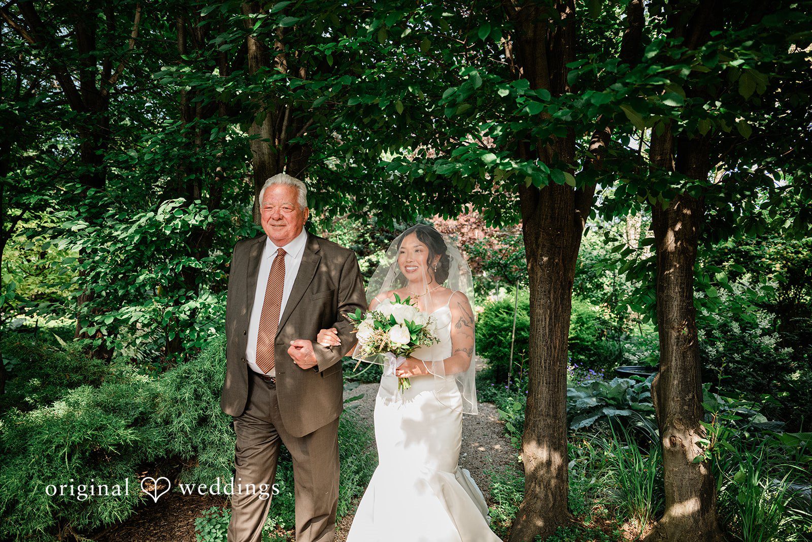 Lauren + Brinton A bride walking alongside a man down a path lined with tall green trees.