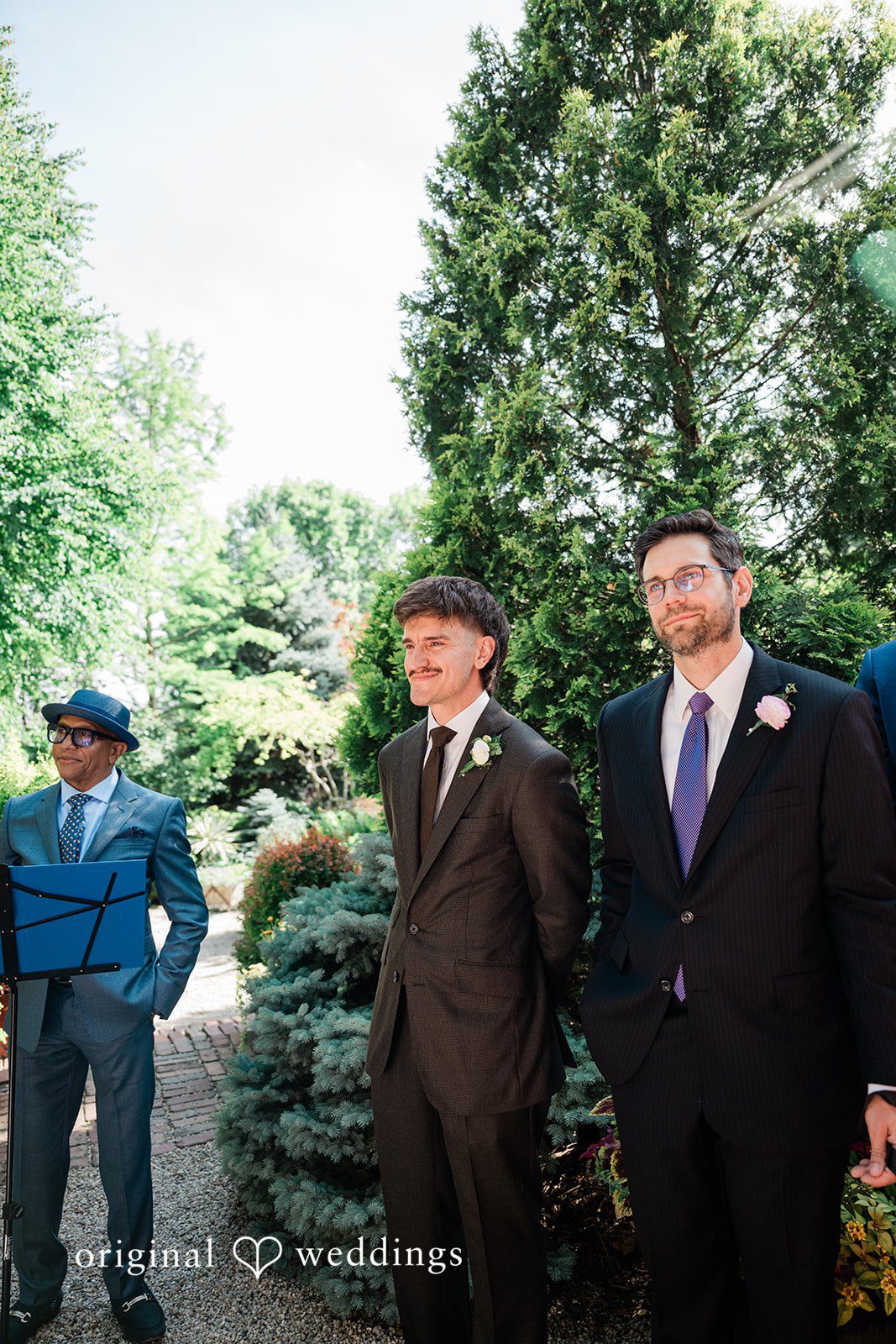 Lauren + Brinton Two men dressed in dark suits standing together in front of greenery.