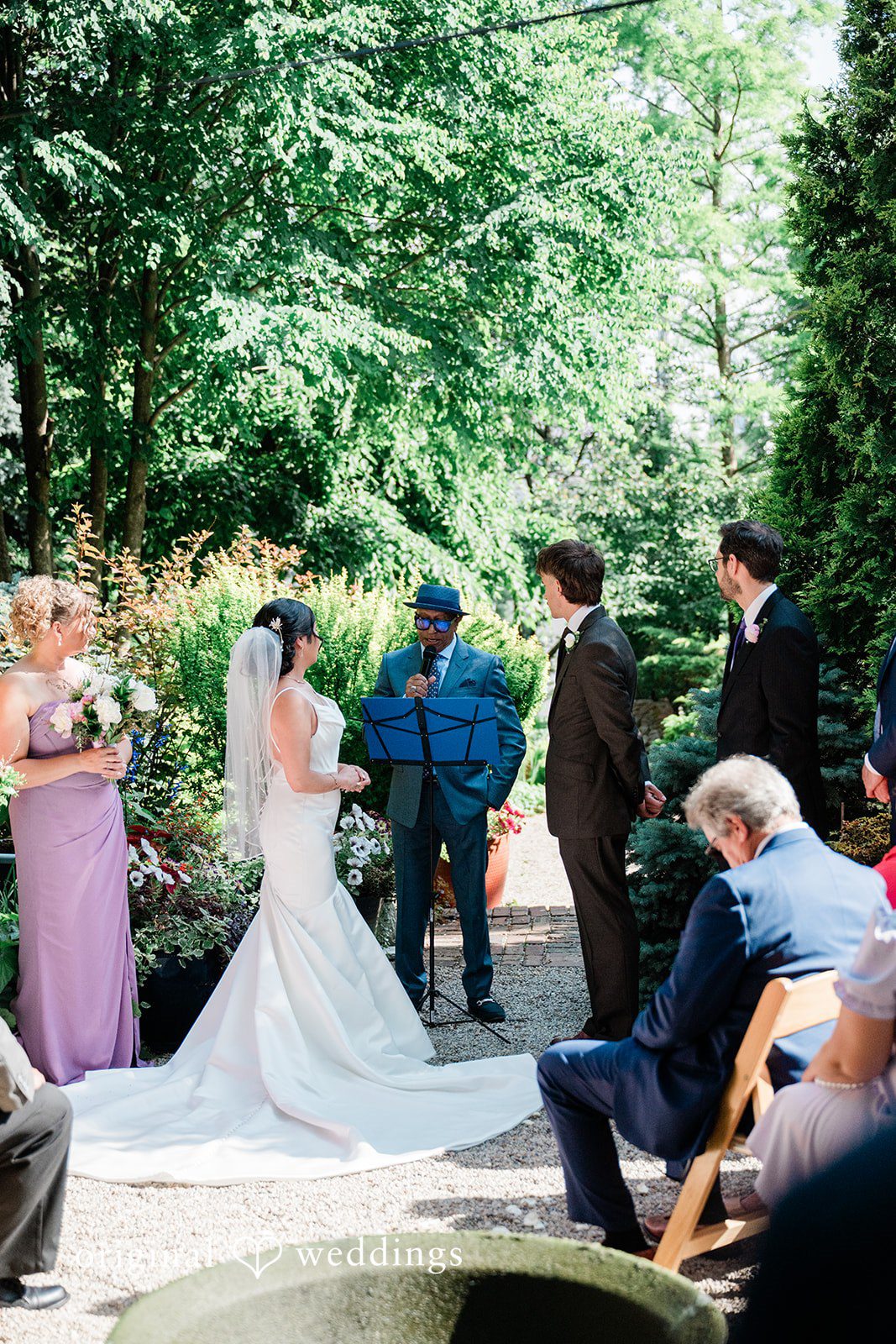Lauren + Brinton A wedding ceremony taking place on a lawn surrounded by trees and guests.