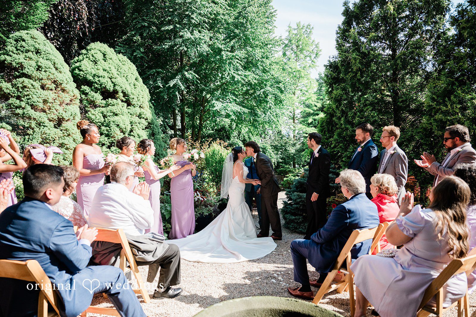 Lauren + Brinton A couple shared first kiss and wedding crowd seated on chairs watching a ceremony in a garden.