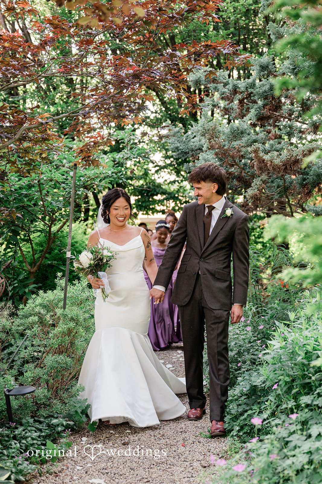 Lauren + Brinton A bride and groom walking hand-in-hand down a scenic wooded path.