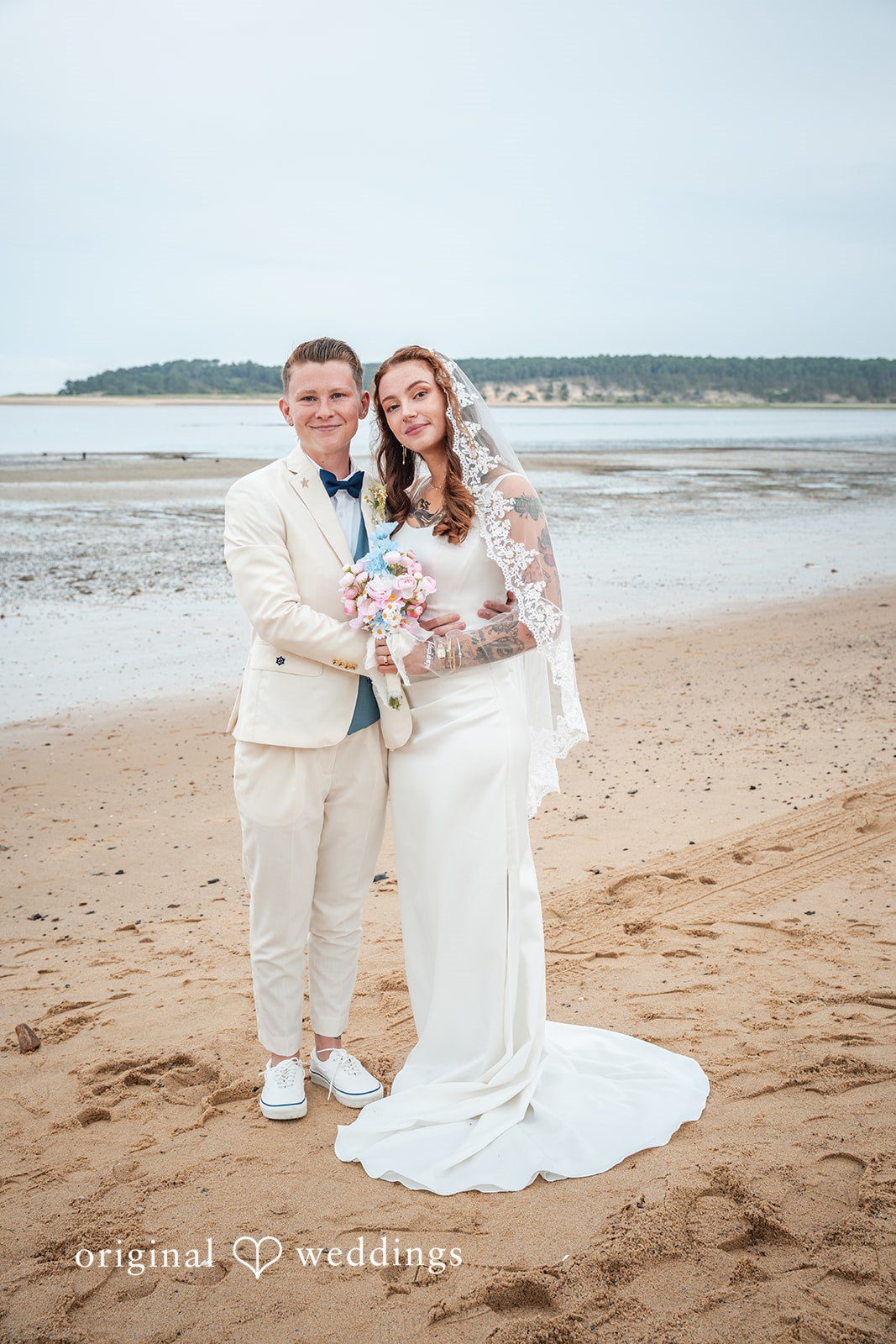 Jacqueline + Sarah A newlywed couple standing together and posing on a wide sandy beach.