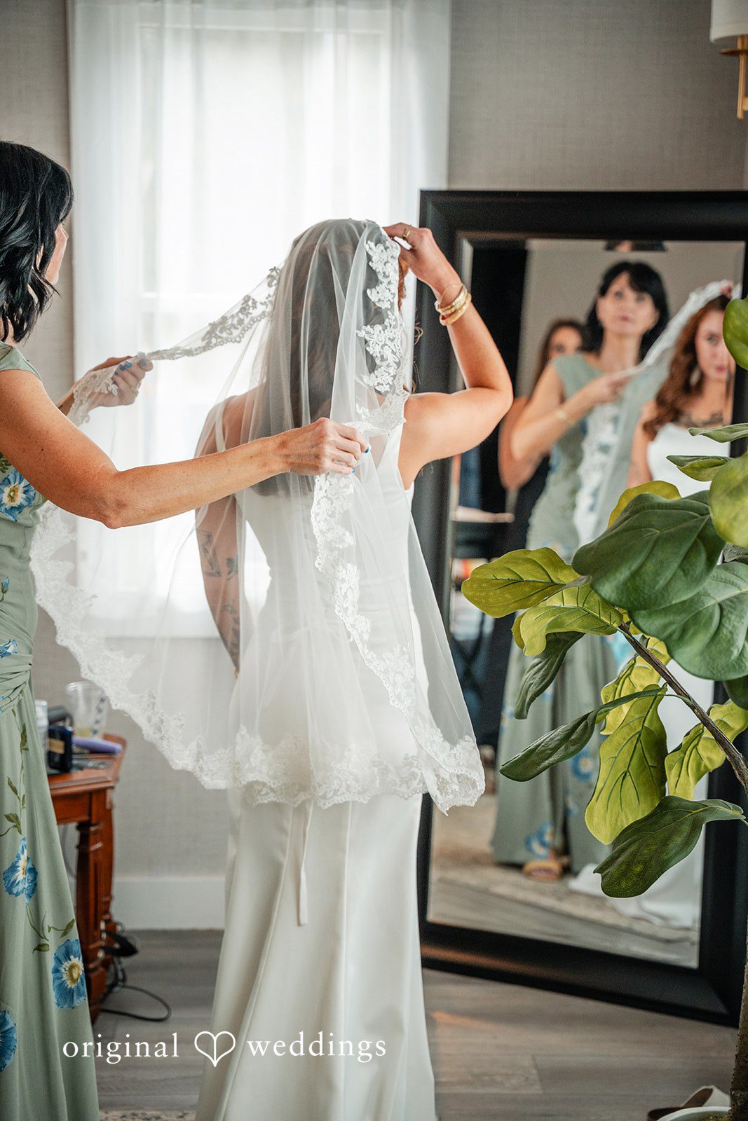 Jacqueline + Sarah A bride looking at herself in a mirror while an assistant adjusts her wedding veil.