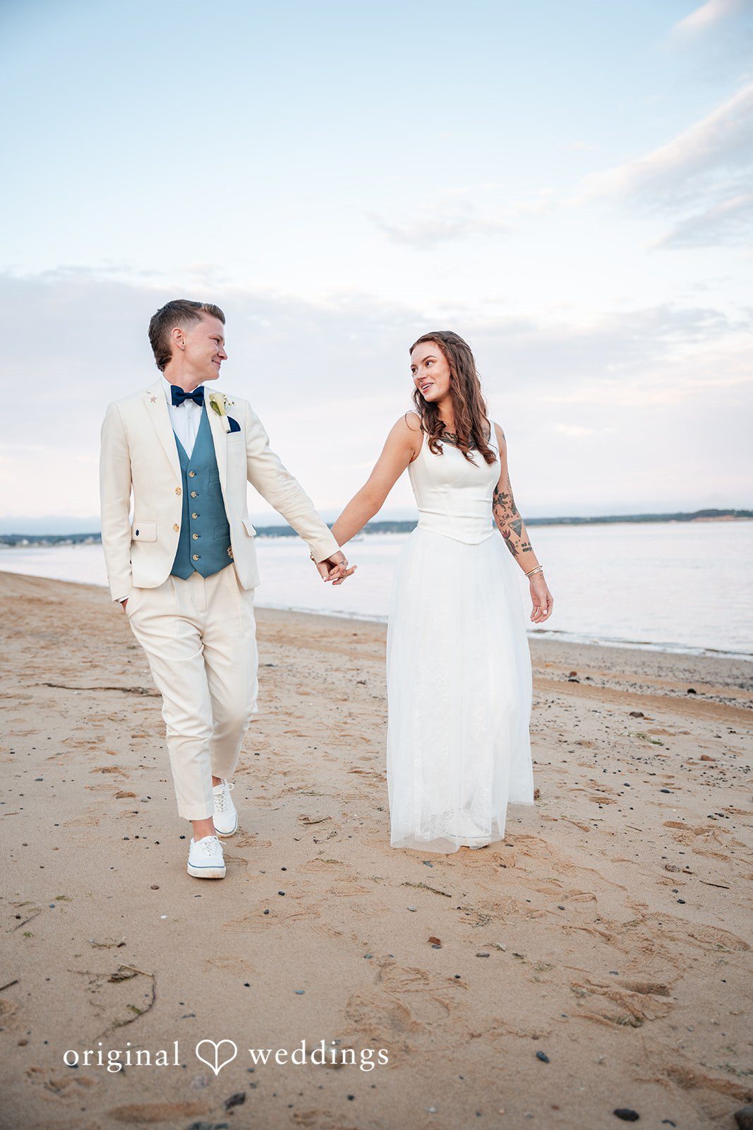 Jacqueline + Sarah A bride and groom holding hands while walking along the shoreline at sunset.
